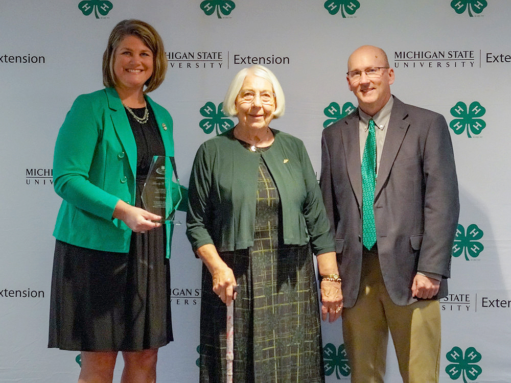 Three individuals at a backdrop. The center person holds an award.
